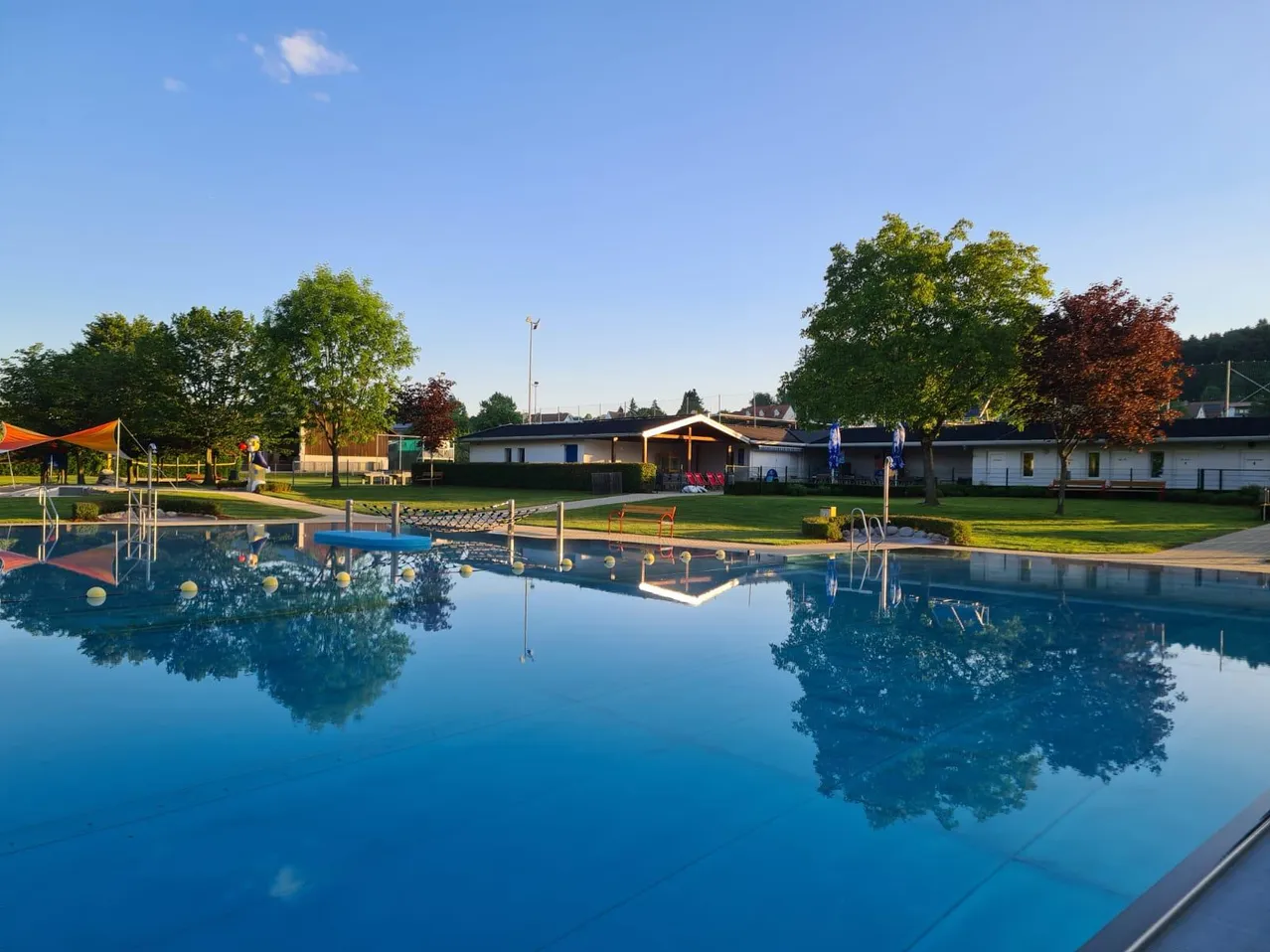 Ein Freibad ist von einem grünen Rasen umgeben. Ein Gebäude steht im Hintergrund, und ein roter Stuhl ist auf dem Rasen platziert. Bäume umgeben den Bereich.