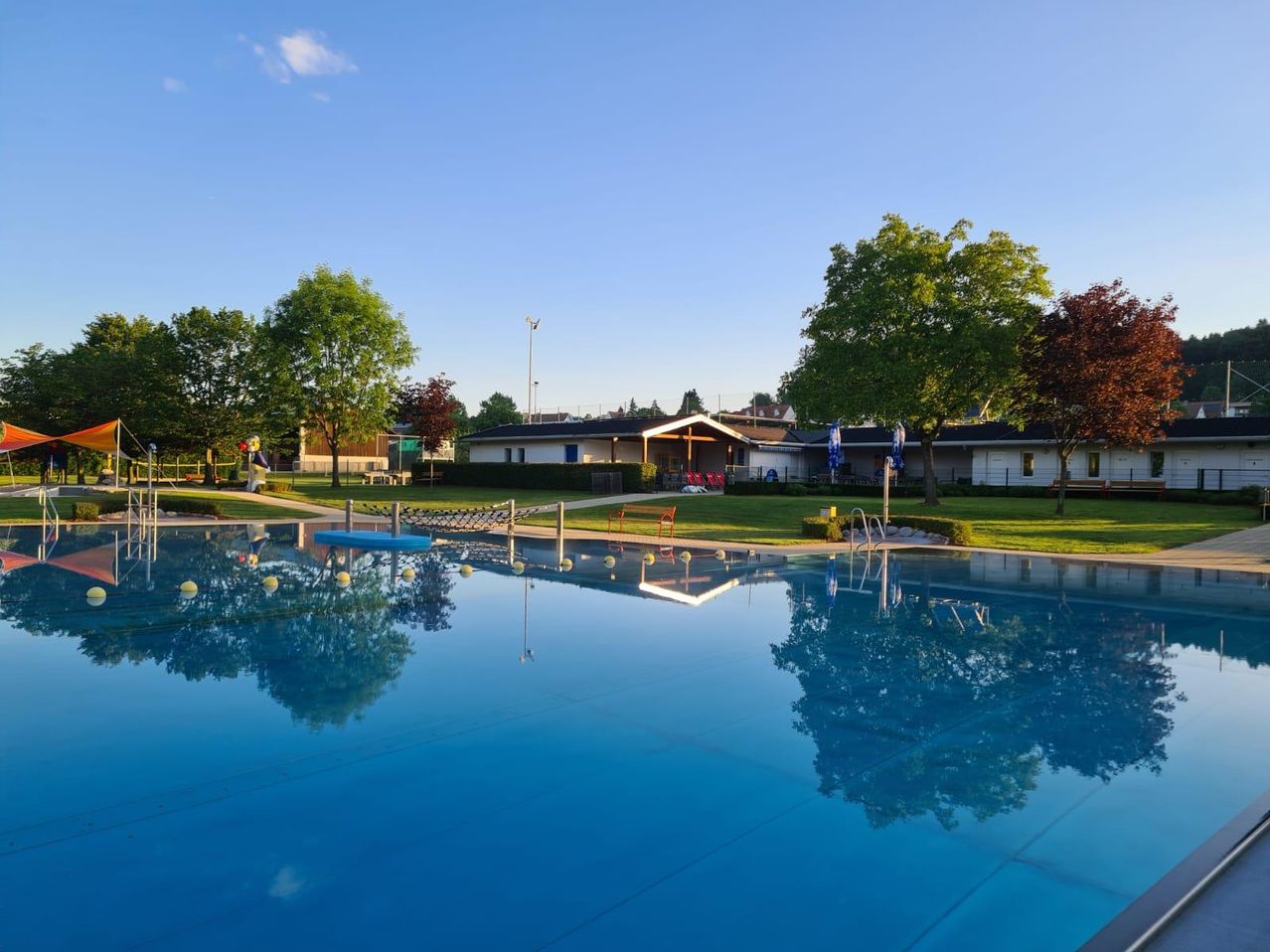 Ein Freibad ist von einem grünen Rasen umgeben. Ein Gebäude steht im Hintergrund, und ein roter Stuhl ist auf dem Rasen platziert. Bäume umgeben den Bereich.