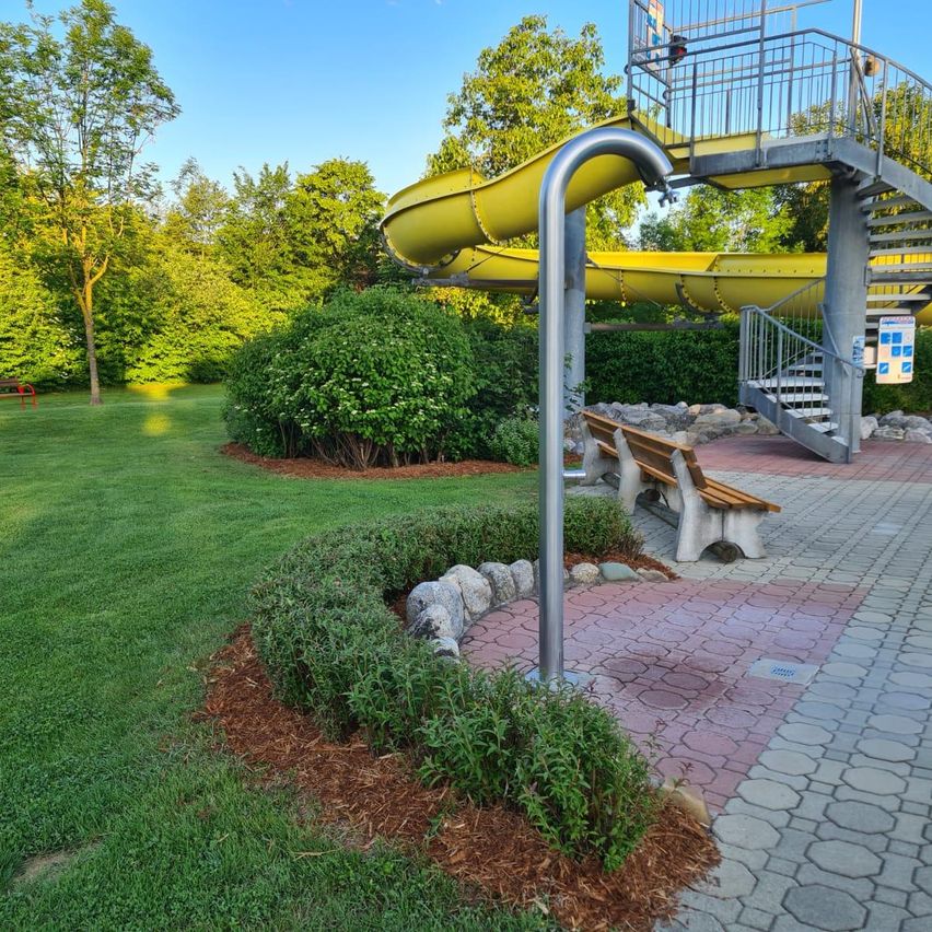 Ein Wasserrutsche im Freien mit einem Metallpfosten. Eine Bank steht neben der Rutsche. Die Rutsche hat eine Treppe mit Handläufen. Es gibt Büsche und Gras am Boden.