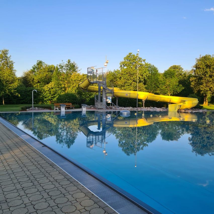 Ein Freibad mit einem gelben Wasserrutsche, umgeben von Bäumen und einer Bank unter einem blauen Himmel.