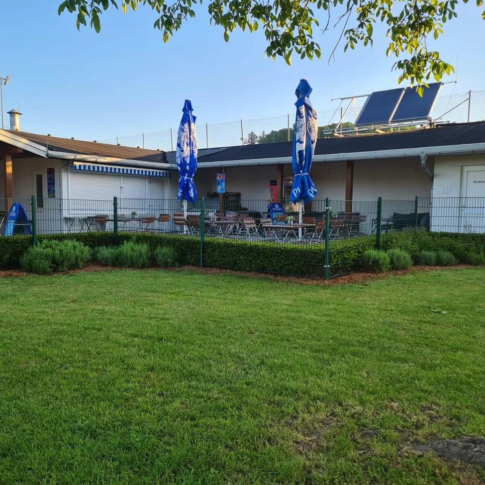 Ein Gebäude mit Solarpanelen auf dem Dach und blauen Sonnenschirmen vor einem Restaurant, mit einem Rasen und einem Zaun.