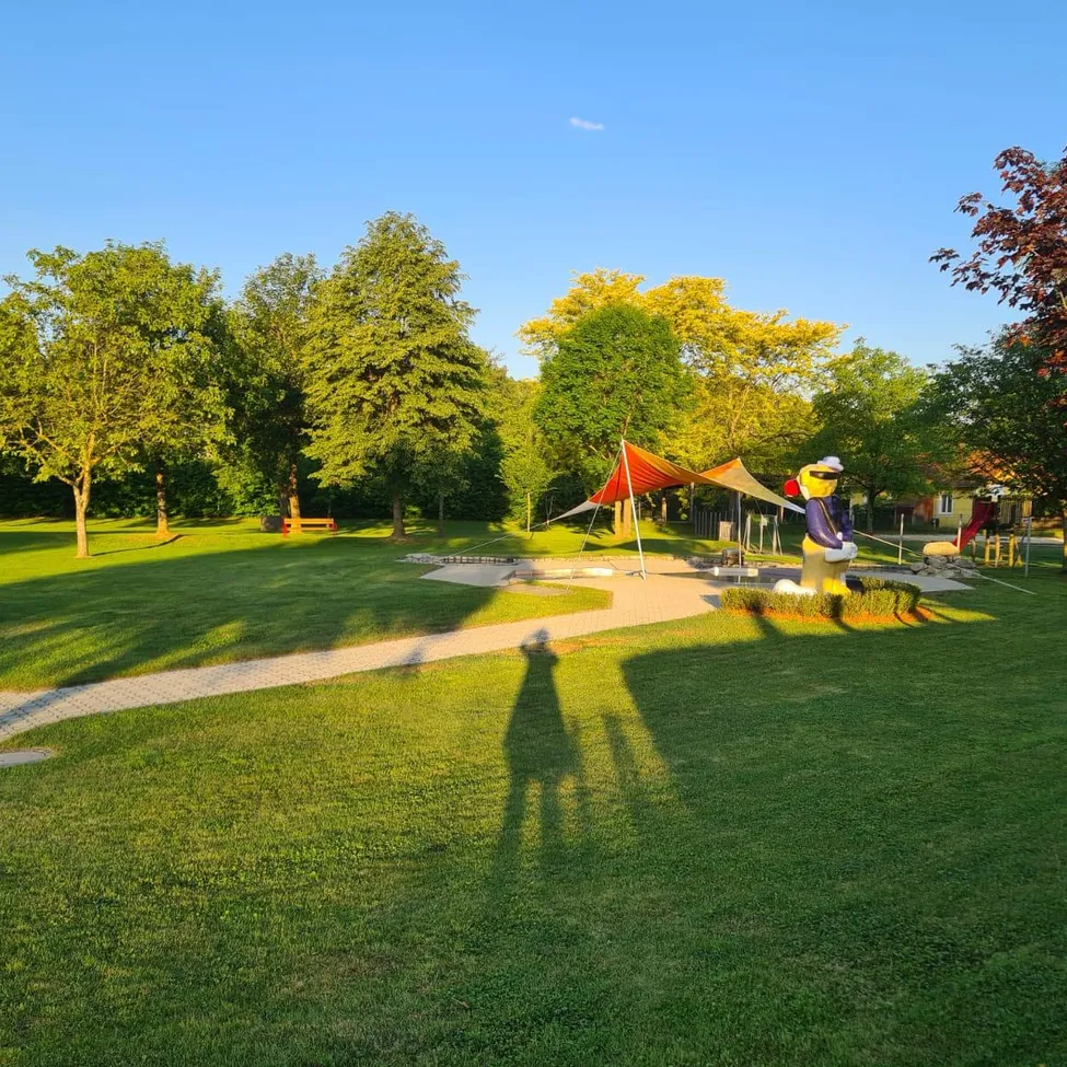 Ein breiter, sonniger Park mit Bäumen, einer gelben Statue und einem roten Baldachin. Schatten fallen auf den Rasen. Eine Bank und eine Rutsche sind in der Ferne.