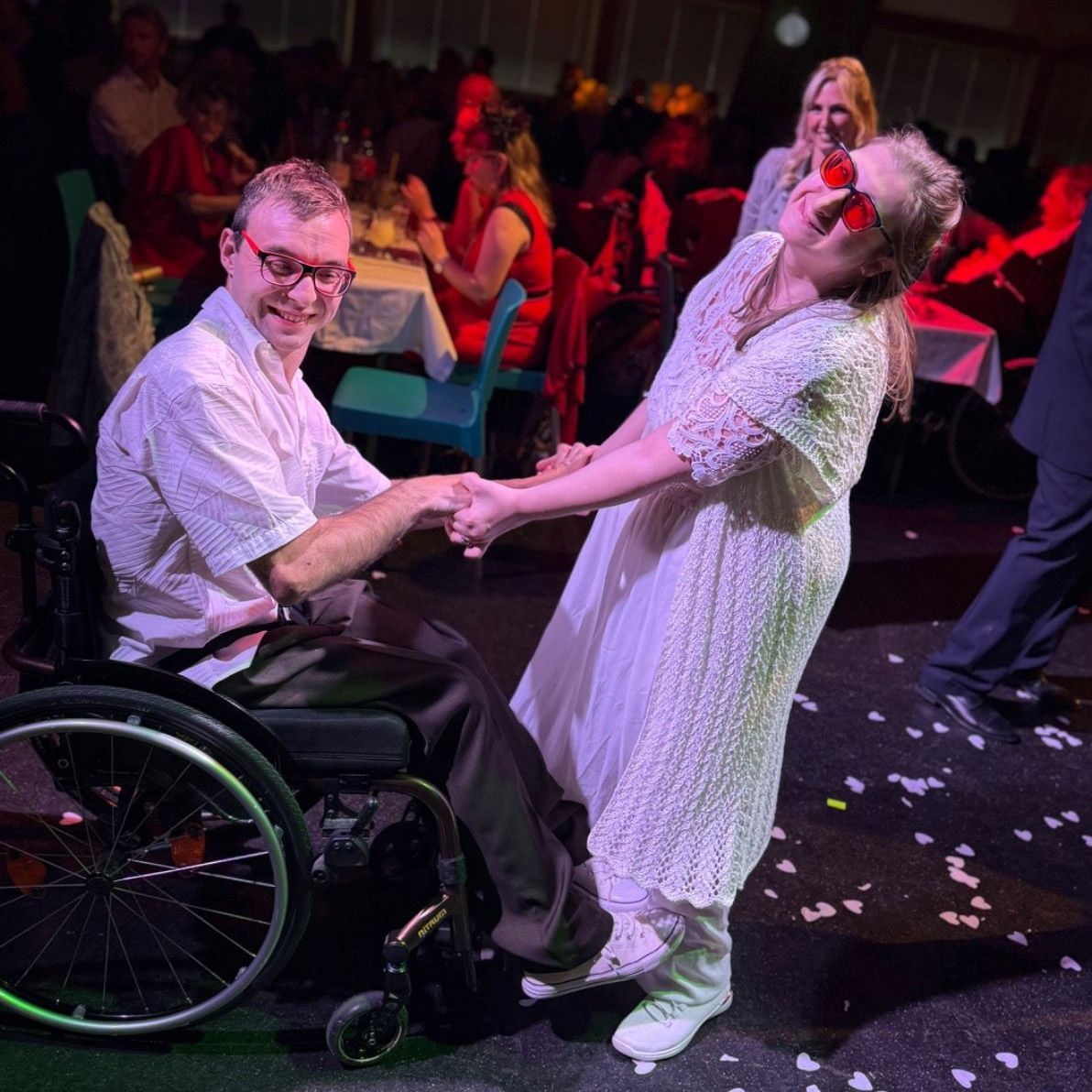 Ein Mann im Rollstuhl und eine Frau in einem weißen Kleid halten Händchen und lächeln. Sie sind von Menschen bei einer Party umgeben.