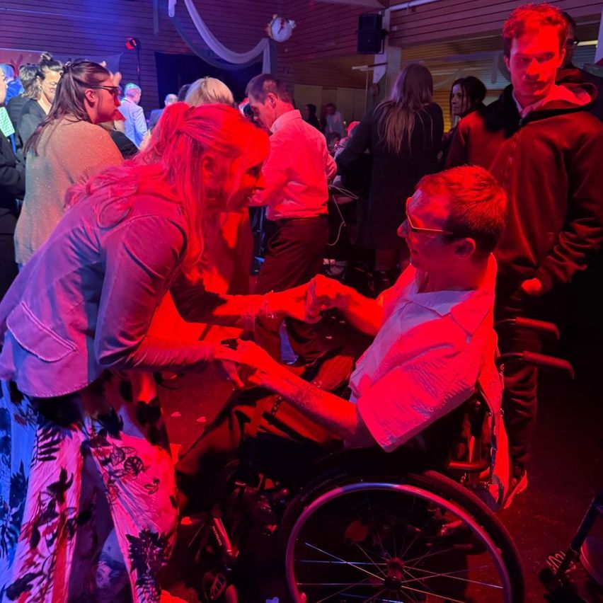 Ein Mann im Rollstuhl unterhält sich mit einer Frau bei einer gesellschaftlichen Veranstaltung. Der Raum ist mit roten Lichtern schwach beleuchtet.