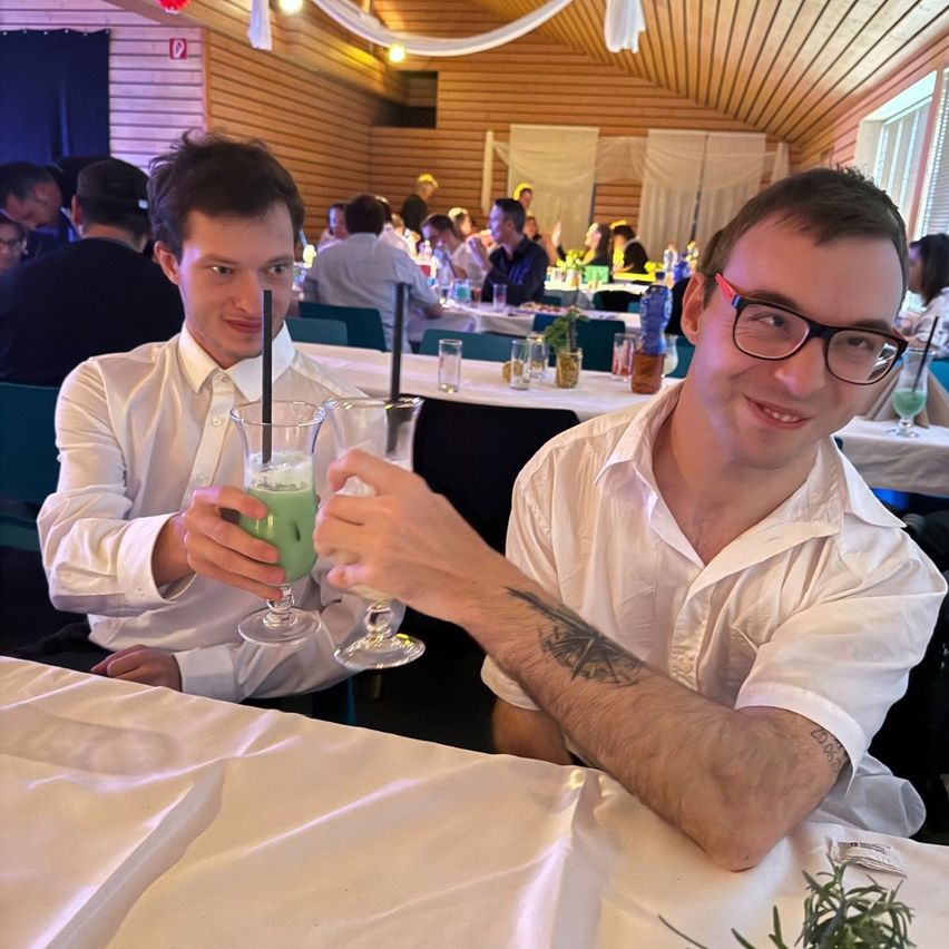 Zwei Männer in weißen Hemden sitzen an einem Tisch, einer hält ein grünes Getränk und lächelt. Hinter ihnen sitzen andere Menschen an Tischen, und die Umgebung scheint eine gesellige Zusammenkunft zu sein.