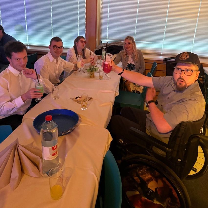 Eine Gruppe von Menschen, einige mit Brille, sitzt um einen Tisch herum und hebt Getränke. Eine Person ist im Rollstuhl. Es gibt Gläser, eine Flasche und eine blaue Tablett auf dem Tisch. Der Hintergrund zeigt ein Fenster mit Jalousien.