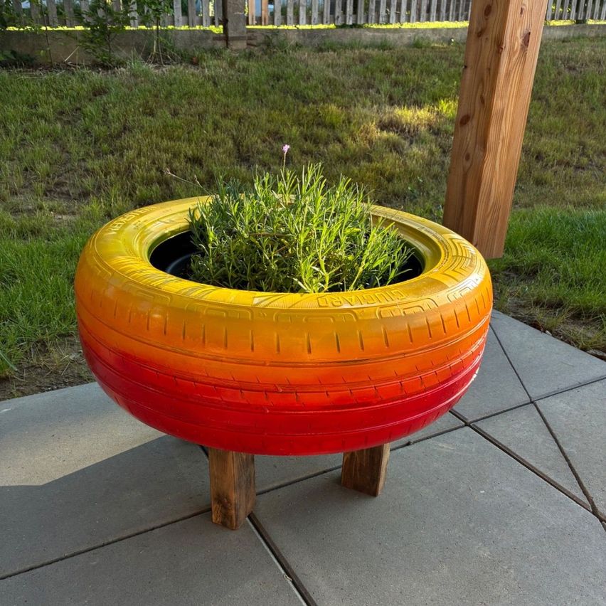 Ein farbenfroher Reifenübertopf mit einer kleinen Kräuterpflanze steht auf einem Holzständer auf einem Betonpflaster neben einem Grasbereich.