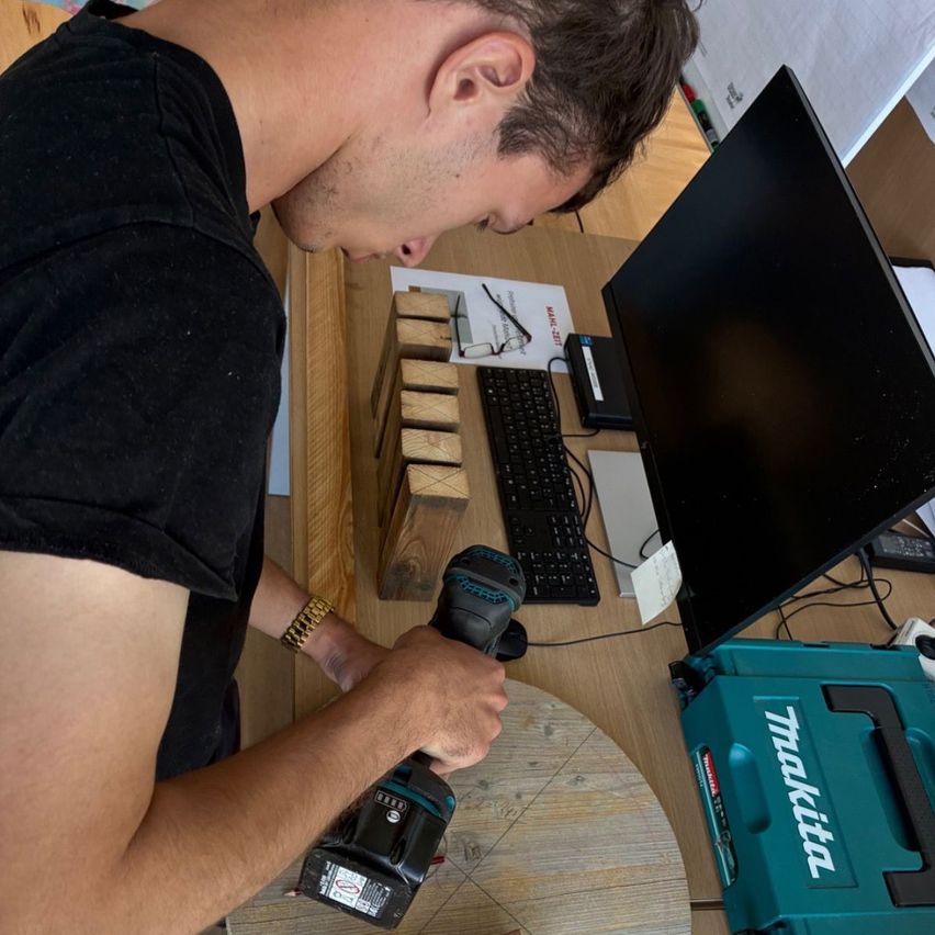 Ein Mann in einem schwarzen T-Shirt benutzt eine Bohrmaschine an einer Holzplatte, mit einem Monitor, einer Tastatur und einem Werkzeugkasten in der Nähe.