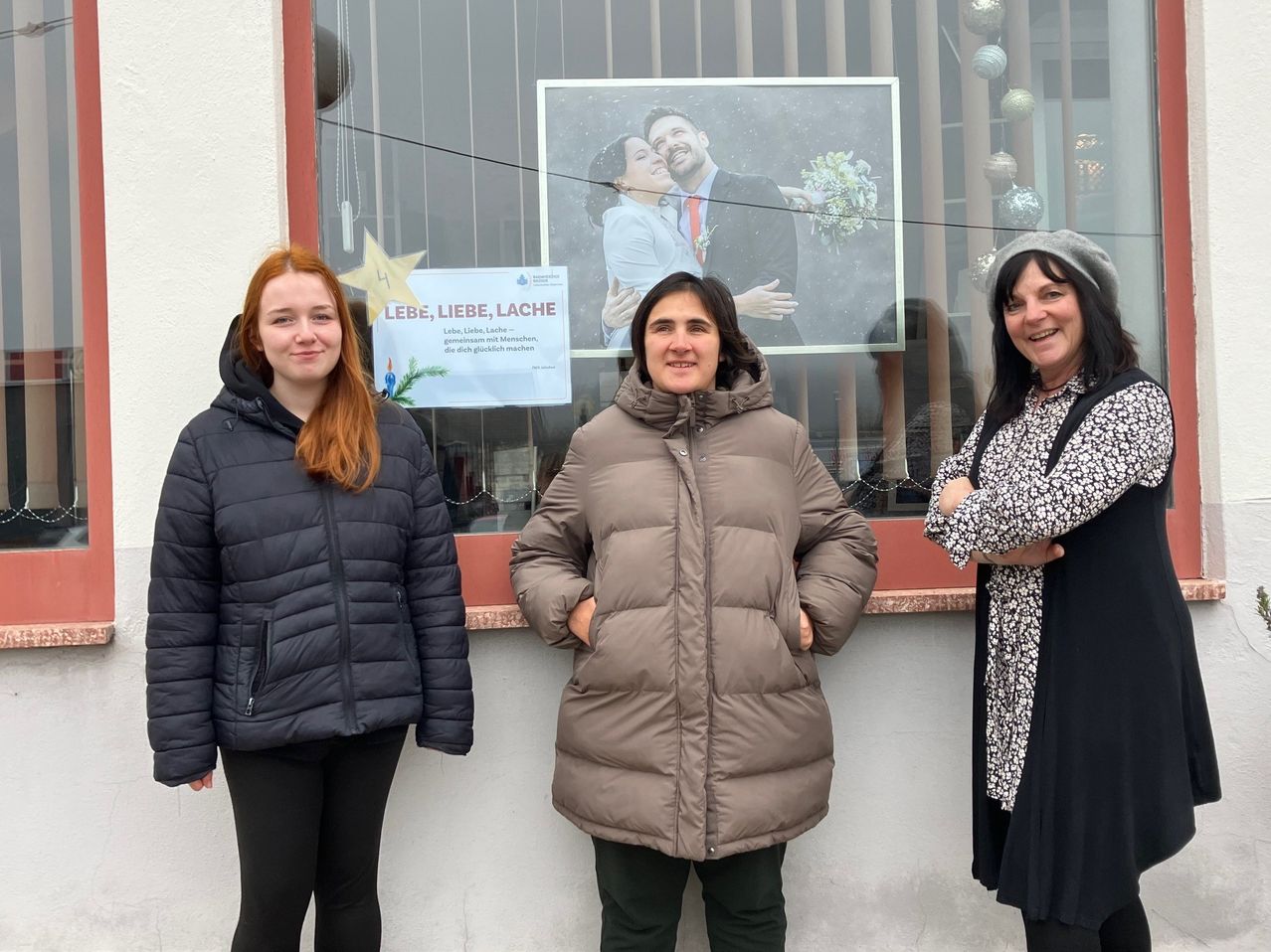 Drei Frauen posieren vor einem Geschäft. Hinter ihnen ist ein Plakat zu sehen, das 'Liebe, Liebe, Lach' besagt. Ein Foto eines Paares ist im Schaufenster zu sehen.