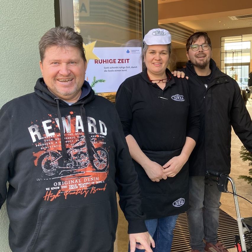 Three people standing outside a building. The man on the left wears a black hoodie with an image of a motorcycle. The woman in the middle smiles, wearing a black apron. The man on the right wears a white cap and glasses. Behind them is a glass door with a yellow star and a sign.