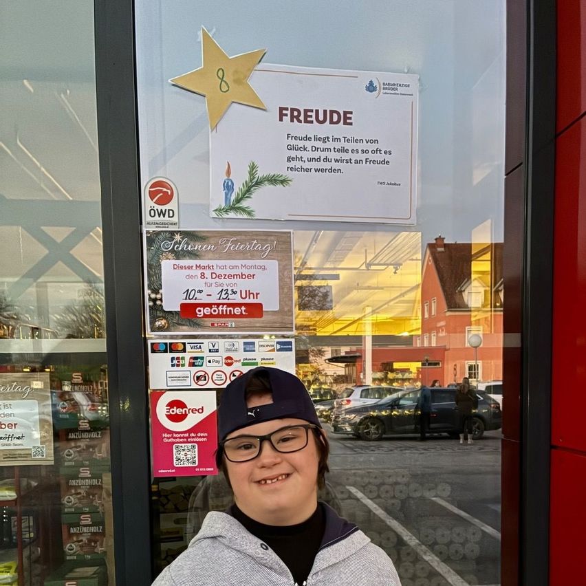 A person stands outside a shop with a door sign reading 'Freude'. Inside, a whiteboard says 'Freude liegt im Teilen von Glück'. A red poster below offers discounts for the holiday season.