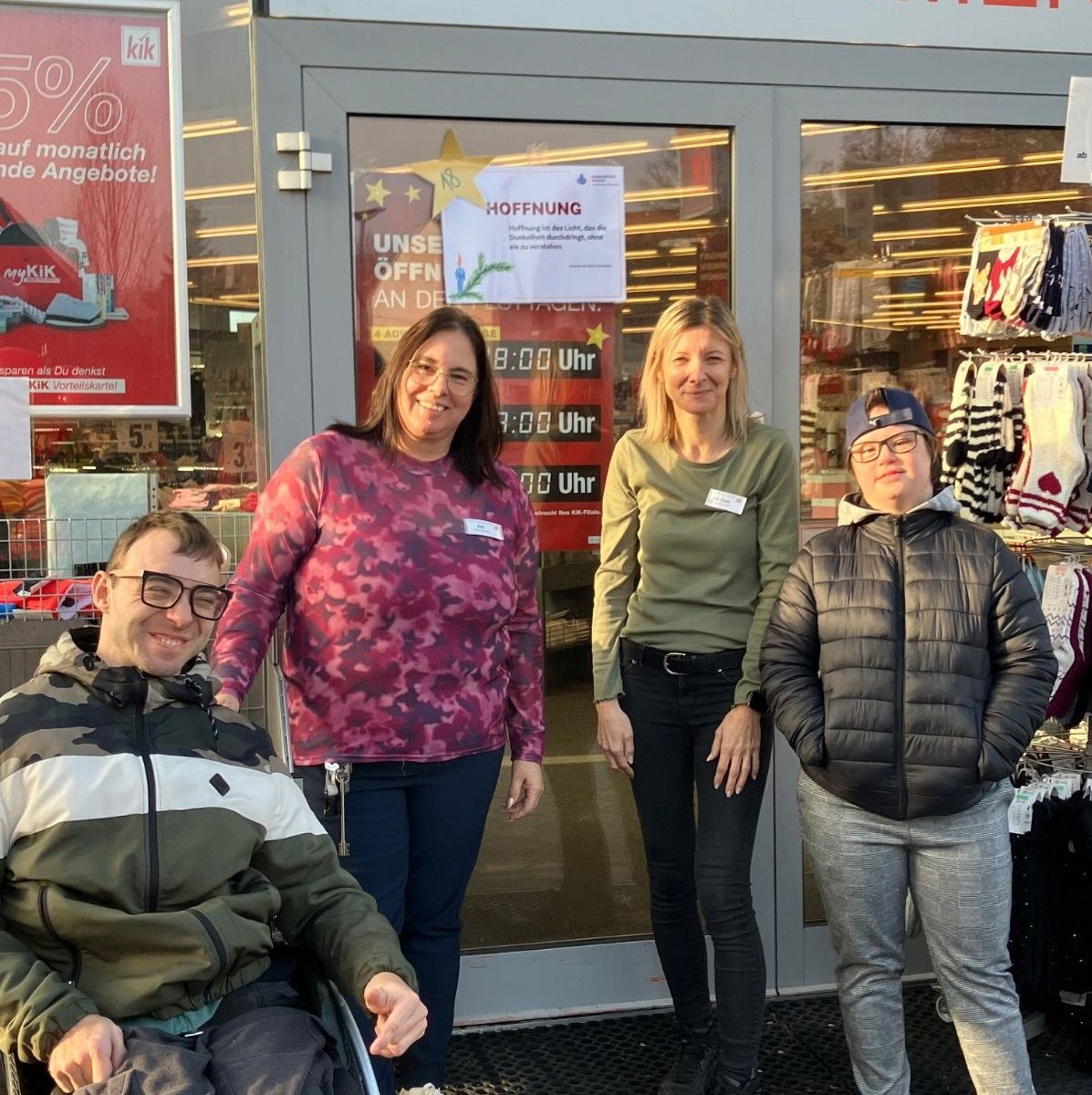 Vier Personen stehen vor einem Geschäft mit einem großen roten Schild. Ein Mann im Rollstuhl lächelt. Eine Frau in einer Camouflage-Bluse steht neben ihm. Eine weitere Frau in Grün steht zwischen zwei Männern.