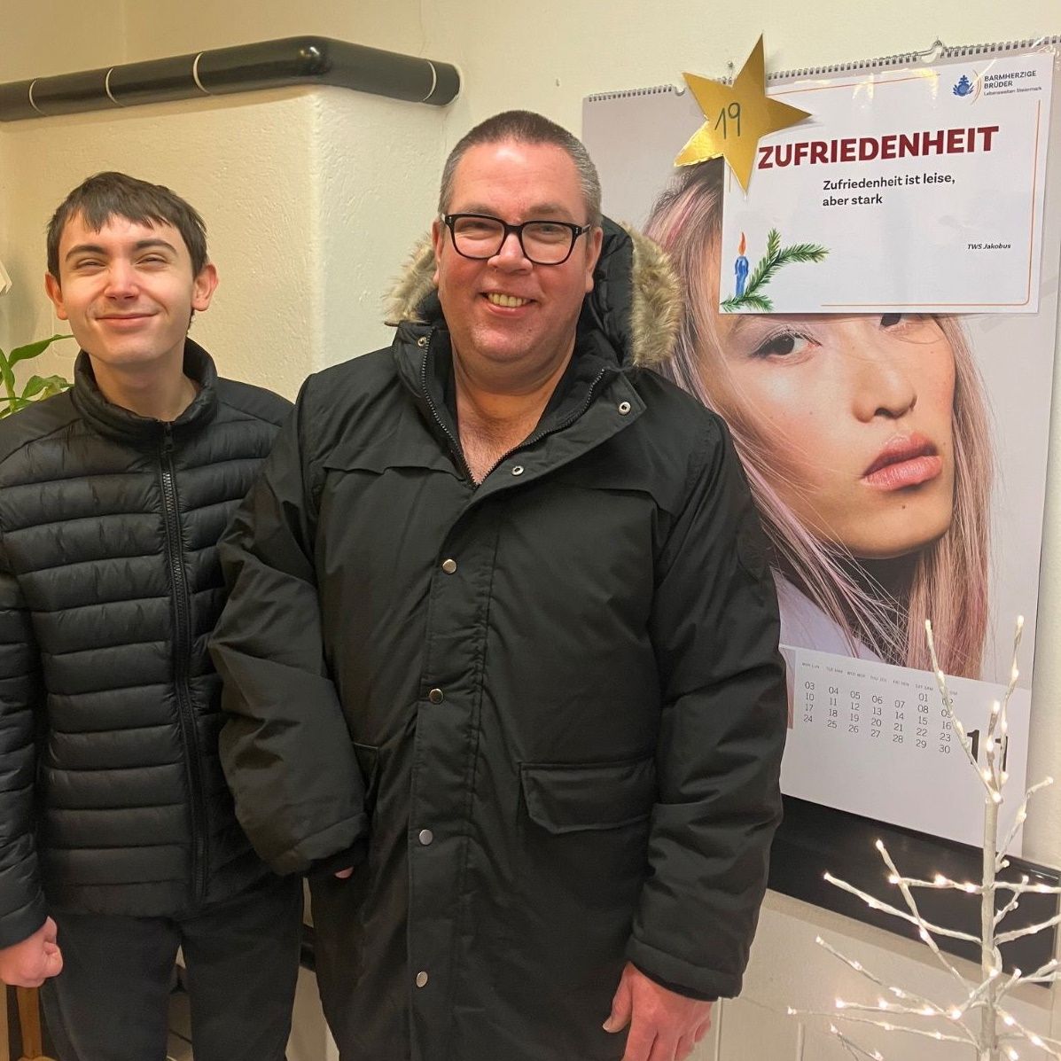 Zwei Männer stehen nebeneinander vor einer Wand mit einem Kalender und einem Baum. Der Mann auf der linken Seite trägt eine gepolsterte Jacke, während der Mann auf der rechten Seite einen Mantel mit Pelzkragen trägt. Sie lächeln beide.