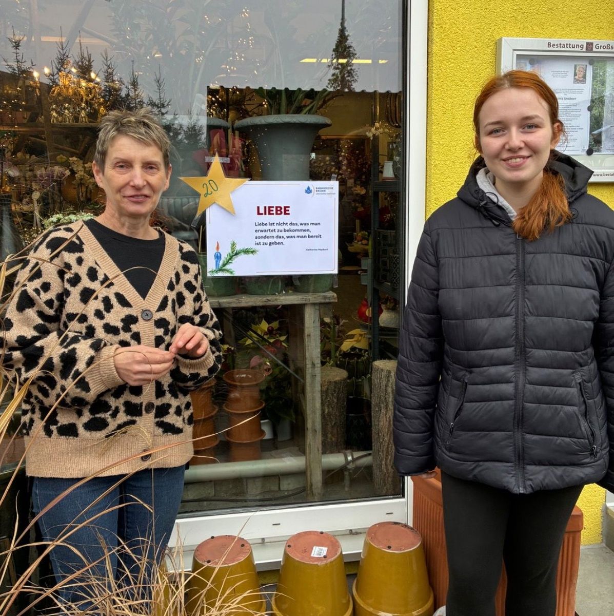 Zwei Frauen stehen vor einem Geschäft, eine trägt einen Leopardenmuster-Pullover, die andere eine schwarze Daunenjacke. Ein gelbes Schild mit weißem Text steht über einem Fenster, das Topfpflanzen zeigt.