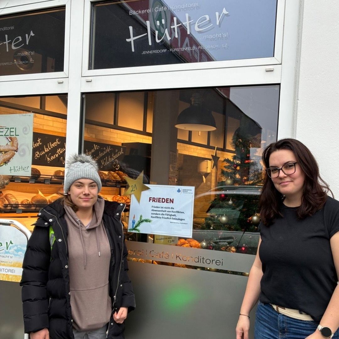 Zwei Frauen stehen vor einer Bäckerei namens Hutter. Eine trägt eine schwarze Daunenjacke, die andere ein schwarzes T-Shirt. Ein Schild zeigt 'Frieden' an und die Bäckerei ist mit Weihnachtsdekorationen geschmückt.