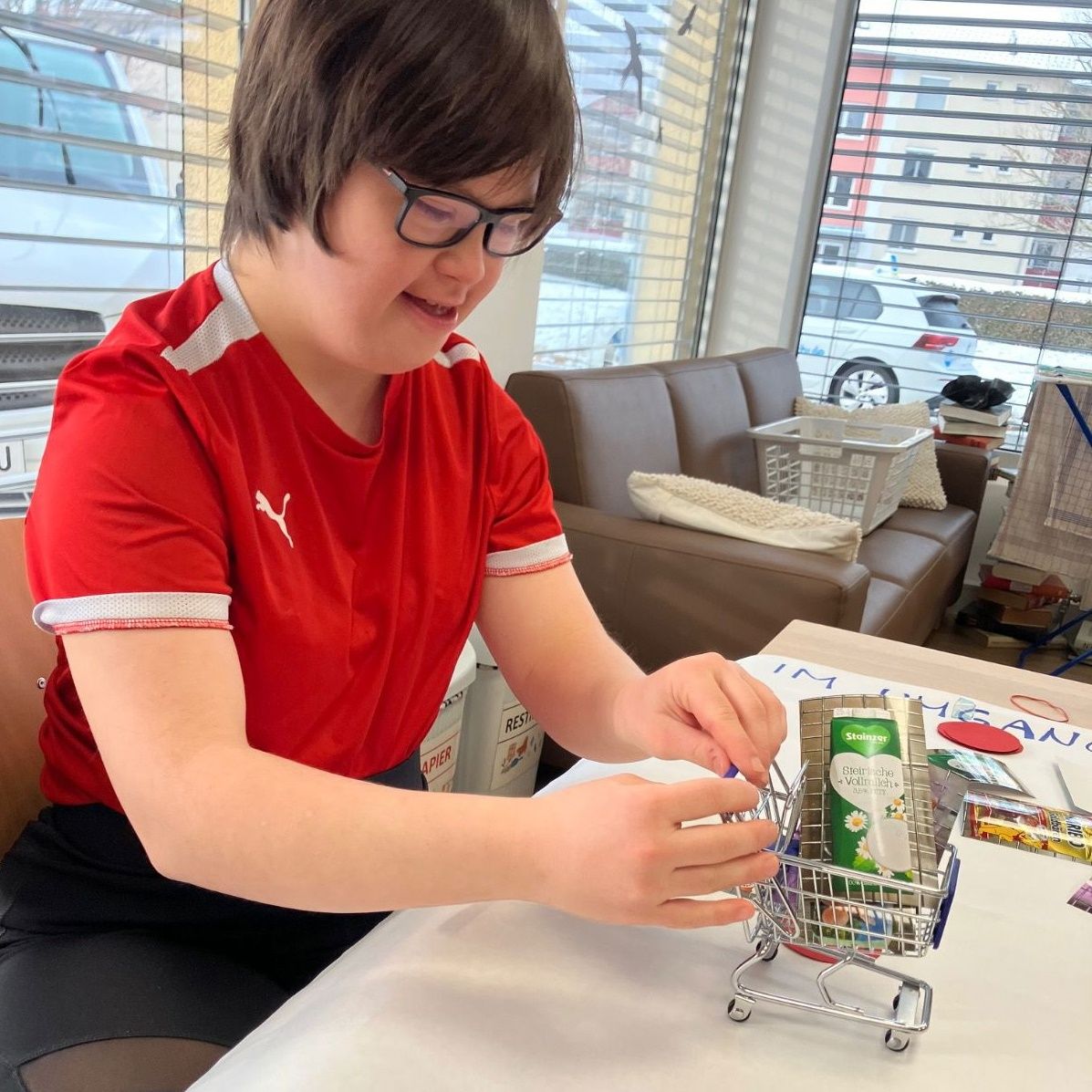 A young person in a red shirt and glasses sits at a table and plays with a toy shopping cart.