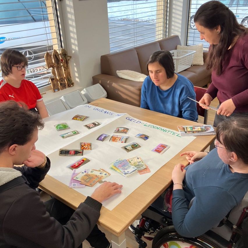 Five individuals are seated around a table covered with cards and money. One individual is in a wheelchair.
