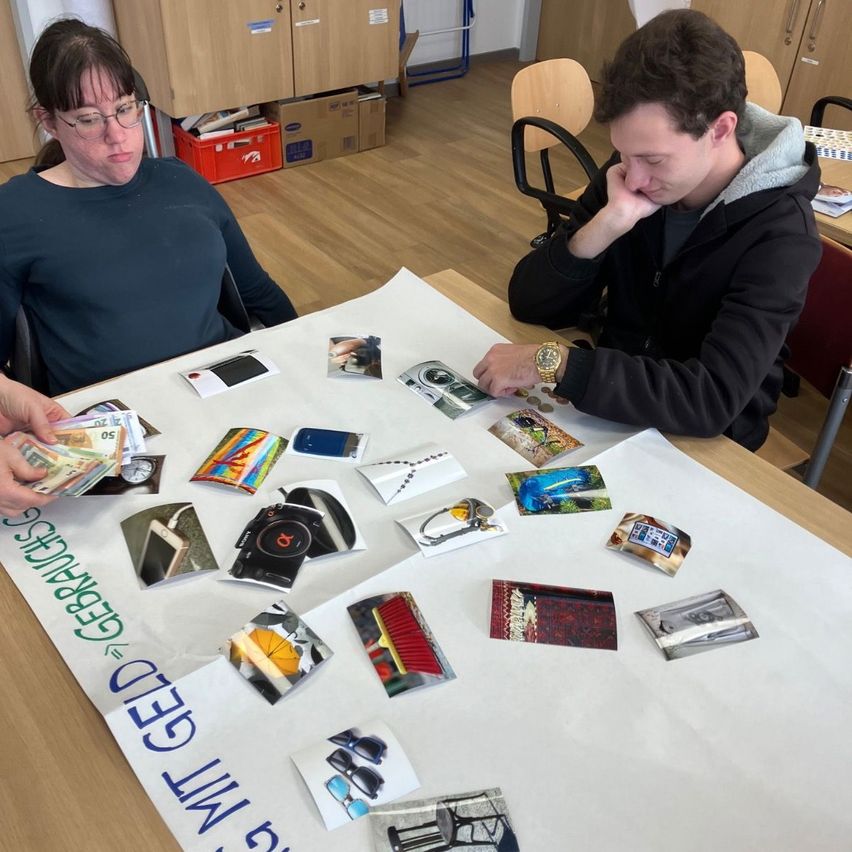 Two individuals, a woman and a man, are sitting at a table. The table has various items like pictures, coins, and a white cloth with 'GEBRAUCHT GEL' written on it. The man is looking at the items on the table while the woman looks away.