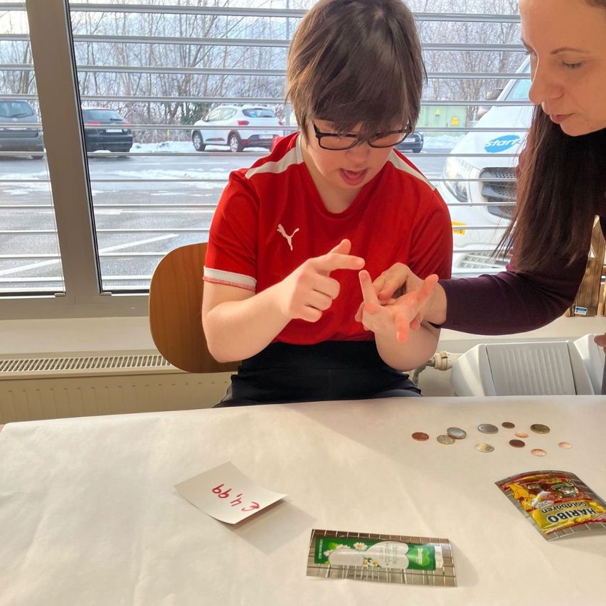 A boy in a red Puma shirt sits at a table, counting coins with a woman, outside a window showing a snowy parking lot.