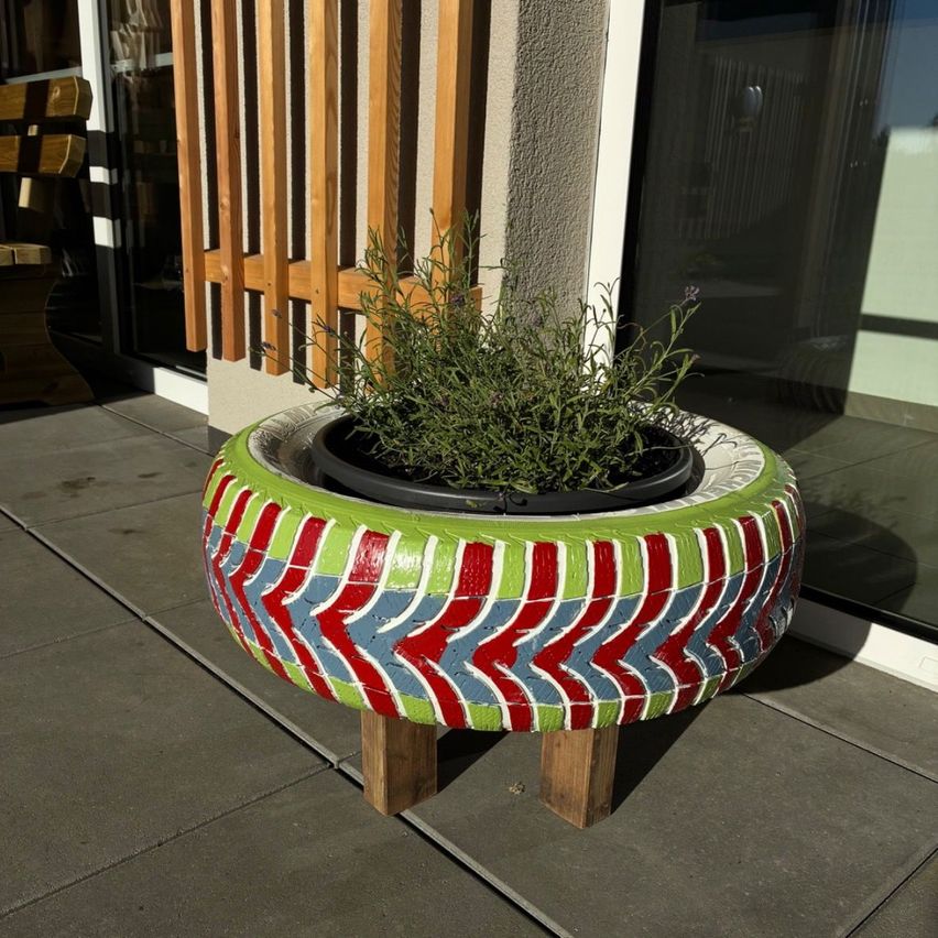 Ein farbenfroher Pflanztopf aus einem recycelten Reifen steht auf einem Holzständer draußen. Der Topf hat eine grüne Pflanze darin.