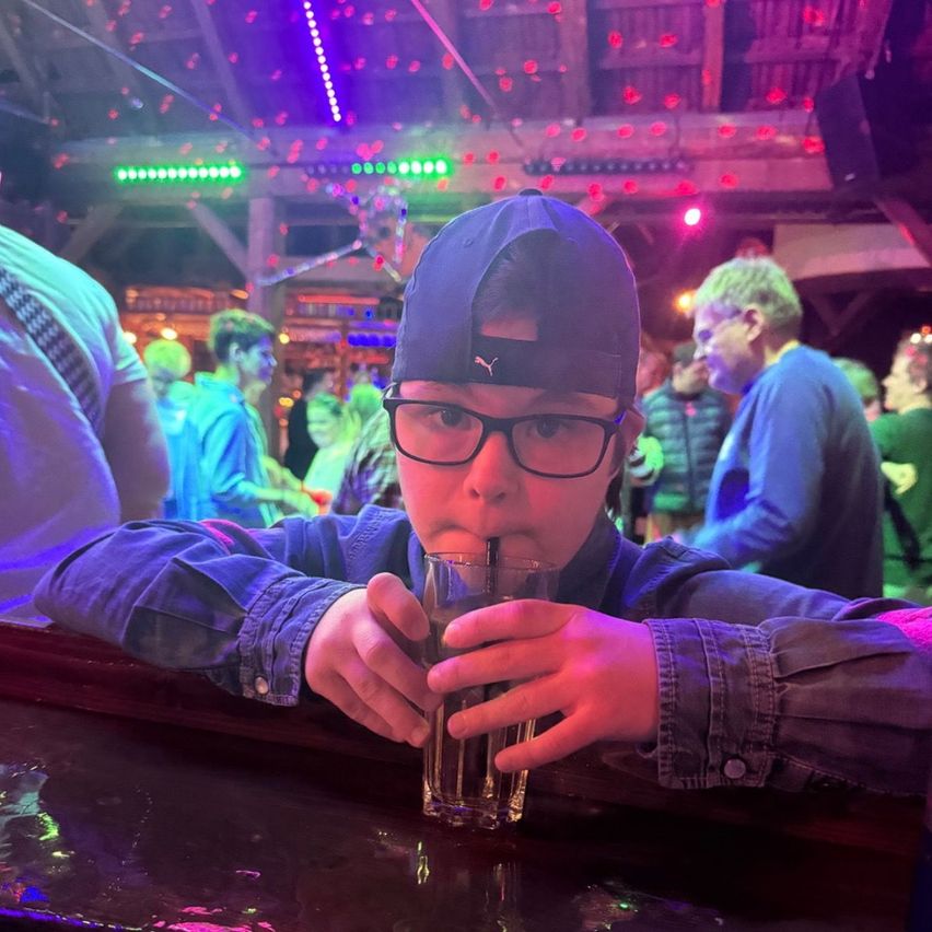 Ein junger Junge mit Brille und Mütze trinkt Wasser an einer Bar in einem überfüllten, farbenfroh beleuchteten Raum.