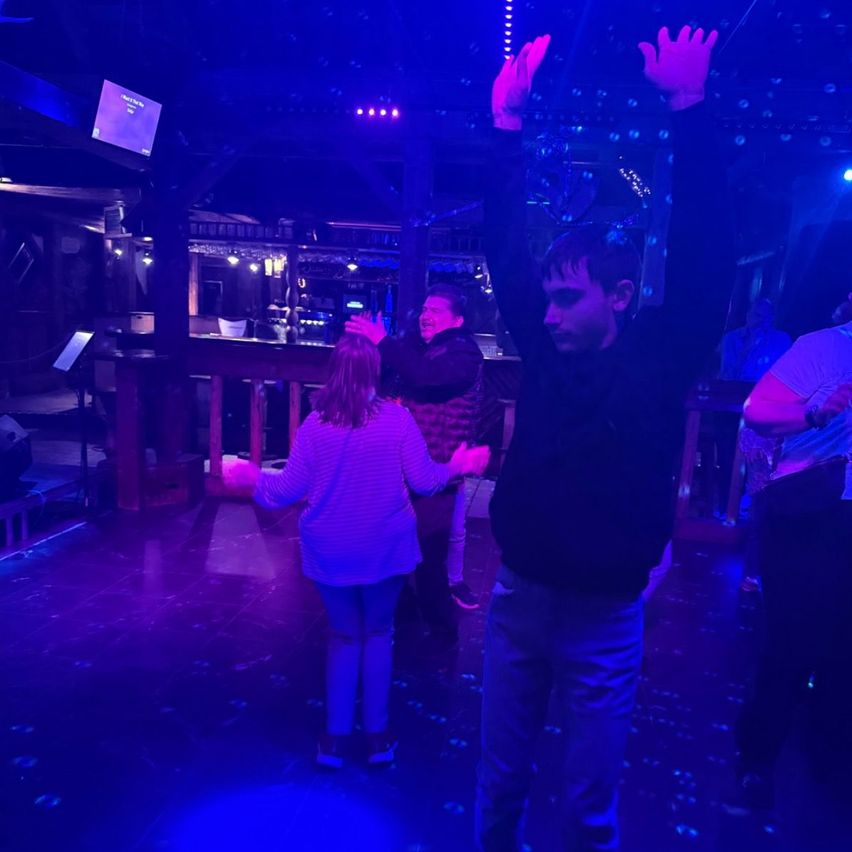 Ein Mann und ein Mädchen tanzen in einem Club mit lila Beleuchtung, ein anderer Mann steht hinter ihnen. Eine Bartheke ist im Hintergrund.