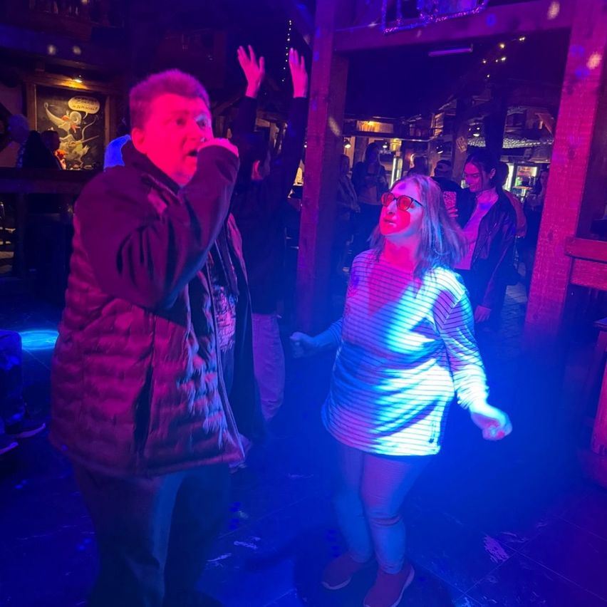 Ein Mann und eine Frau tanzen in einer Bar mit lila und blauen Lichtern. Der Mann hält ein Mikrofon und die Frau trägt eine Brille.