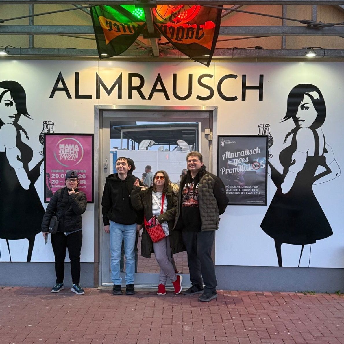 Vier Personen stehen vor einem Geschäft mit dem Namen Almrrausch am Schild. Drei von ihnen lächeln und eine hat eine Tasche. Es gibt zwei Poster mit einer Frau an der Wand.