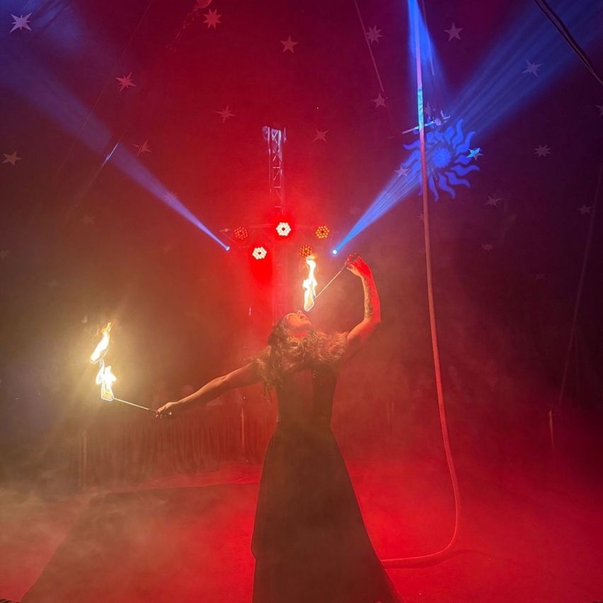 Eine Künstlerin in einem dunklen Kostüm jongliert mit brennenden Stäben unter roten und blauen Lichtern in einem Zirkuszelt.