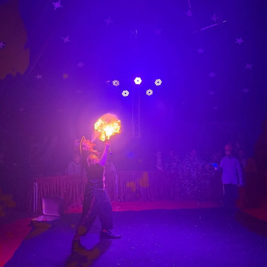 Ein Künstler steht auf der Bühne und pustet Feuer, während eine Menge zusieht, unter einer violett beleuchteten Decke.