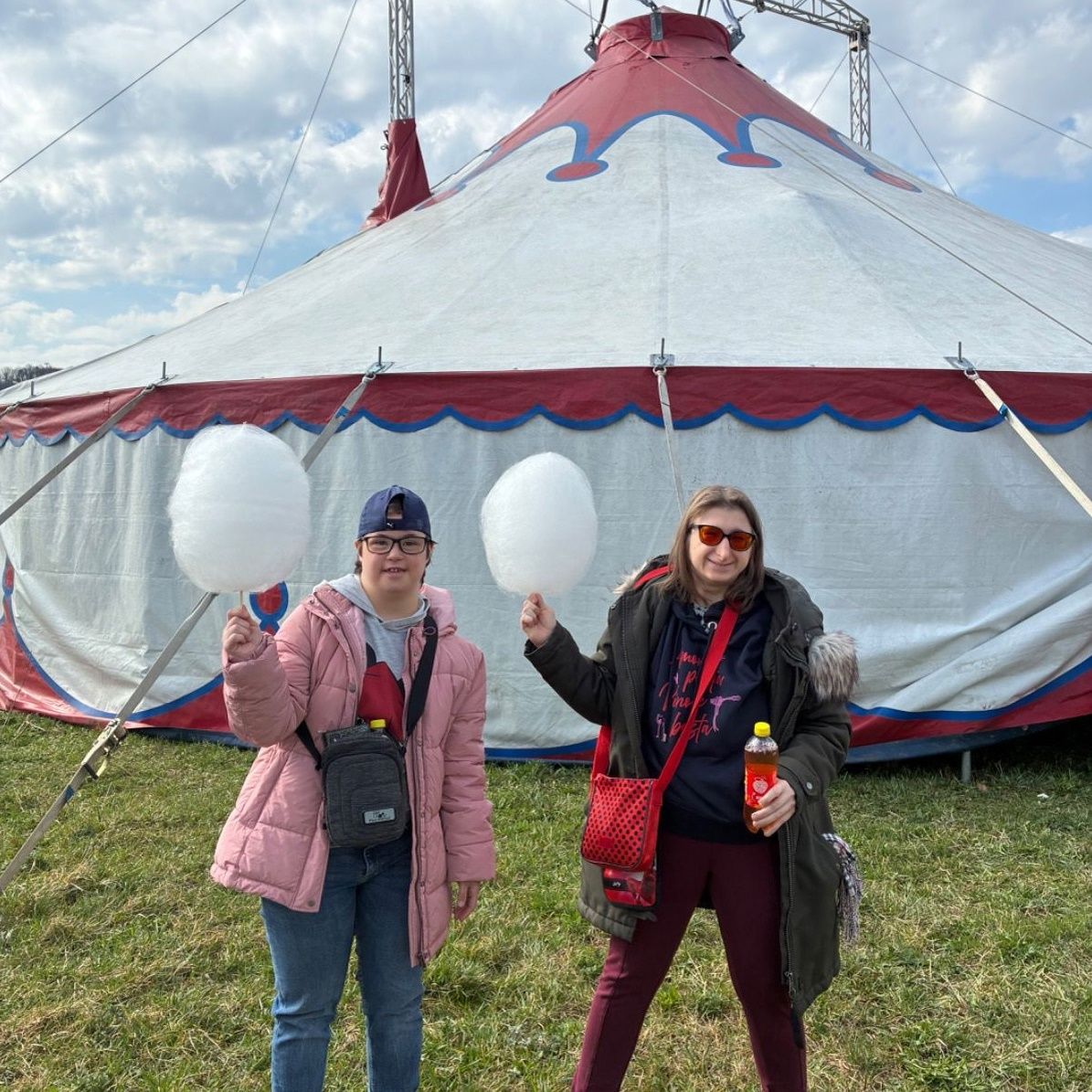 Zwei Frauen stehen vor einem Zirkuszelt und halten Zuckerwatte, eine mit einer roten Tasche und die andere mit einer Flasche. Das Zelt ist groß mit roten und weißen Streifen.