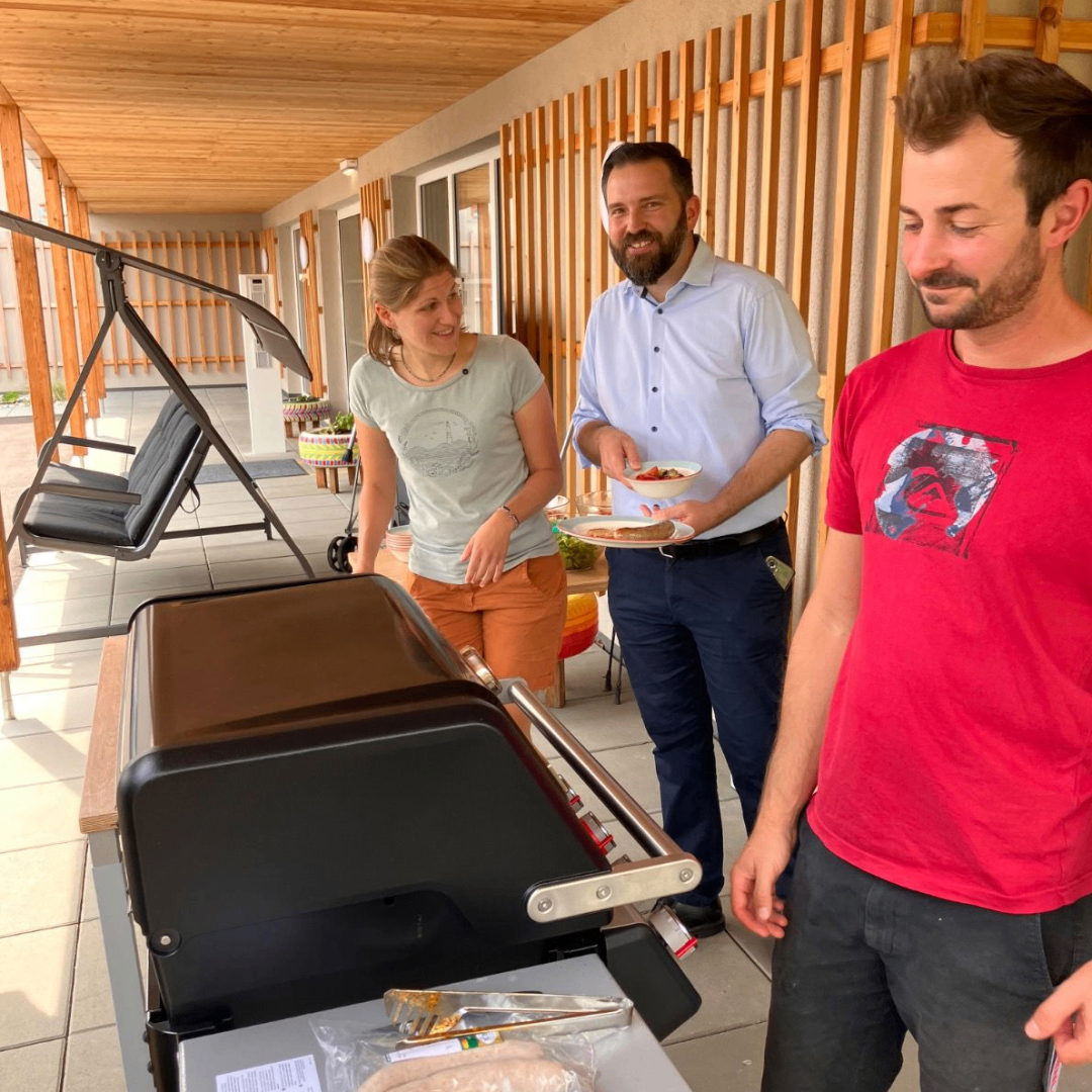 Three people stand around a grill on a patio, one holding plates of food, another smiling, and the third looking at the camera.