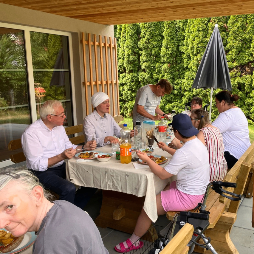 A group of people are sitting around a table in a garden, eating food and drinking. One person is standing and appears to be serving food. There is a wooden fence and trees in the background.