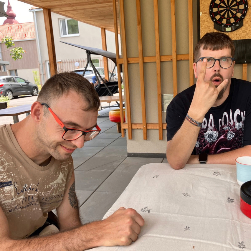 Two men are sitting at a table in an outdoor area, one wearing glasses and a bracelet, the other making a rock sign. Behind them is a dartboard and a building.