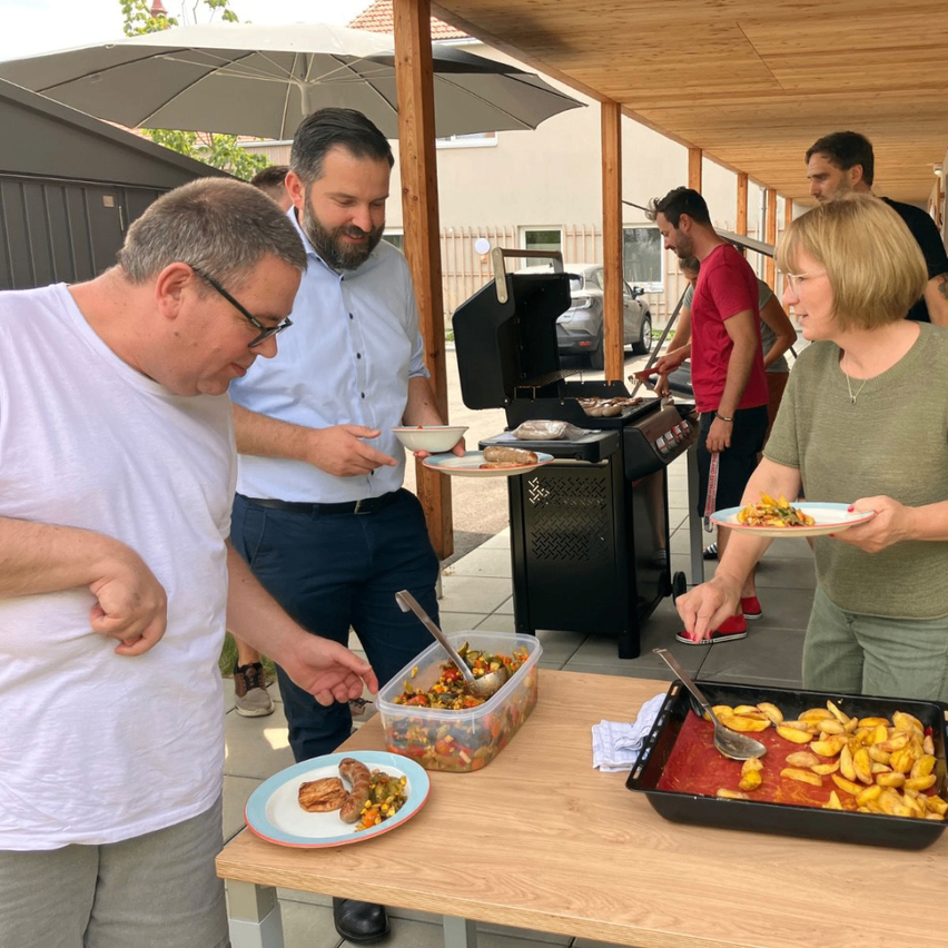 A group of people are enjoying a meal outdoors, with one person grilling food on a black grill. The table is set with plates of food, and others are holding plates.