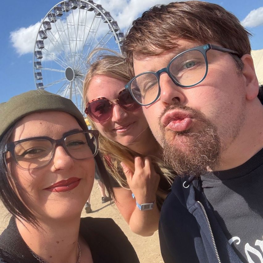 Drei Personen lächeln für die Kamera vor einem Riesenrad. Die Frau links trägt eine Mütze und eine Brille. Die Frau in der Mitte trägt eine Sonnenbrille. Der Mann rechts trägt eine Brille und einen Bart.