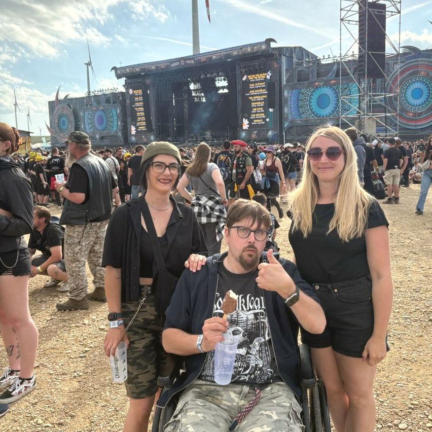 Drei Menschen posieren für ein Foto bei einem Musikfestival, mit einer großen Bühne und einer Menge im Hintergrund. Die Frau auf der rechten Seite lächelt in die Kamera.