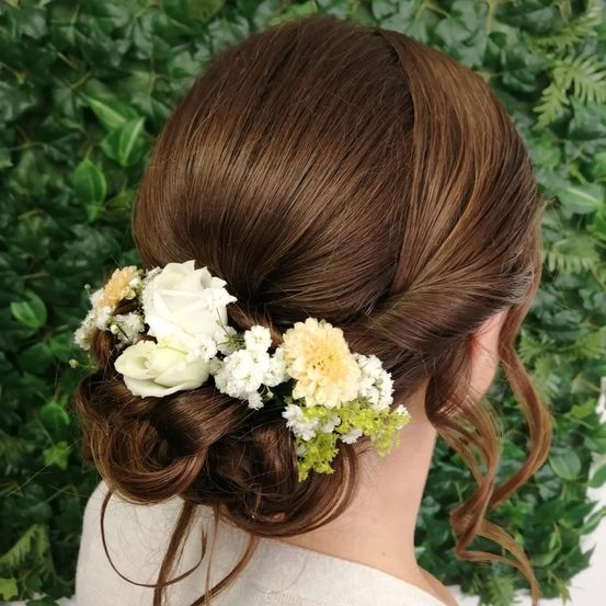 Eine Frau mit braunen Haaren in einer eleganten Hochsteckfrisur, geschmückt mit weißen und gelben Blumen, steht vor einem grünen Hintergrund.