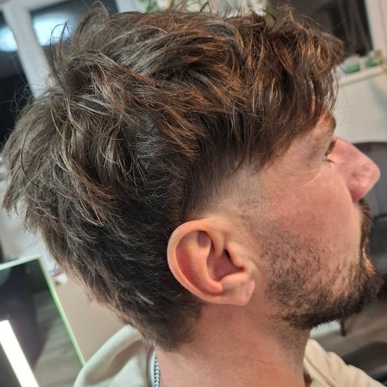 Ein Mann mit kurzen, zerzausten braunen Haaren und Bart schaut zur Seite. Er trägt ein weißes Hemd. Sein linkes Ohr ist sichtbar.