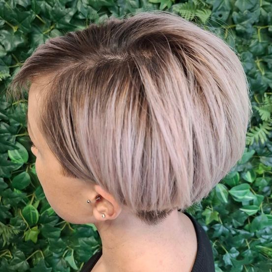 Eine Frau mit kurzen blonden Haaren schaut über ihre Schulter, ihr Profil ist sichtbar. Sie steht vor einer grünen Pflanzenwand.