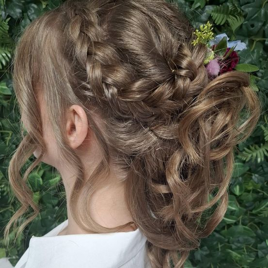 Ein Mädchen mit geflochtener Frisur und Blumenschmuck steht vor einem Hintergrund aus grünen Blättern.