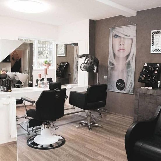 Ein gut beleuchteter Salon mit schwarzen Stühlen und einem weißen Schreibtisch, einem Poster einer Frau an der Wand, einem Spiegel und einer Uhr.