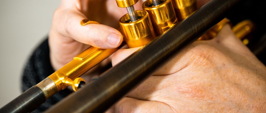 Eine Person hält eine goldene Trompete mit ihren Händen und konzentriert sich auf die Ventile. Der Hintergrund ist unscharf, was auf ein Studio-Setting hindeutet.