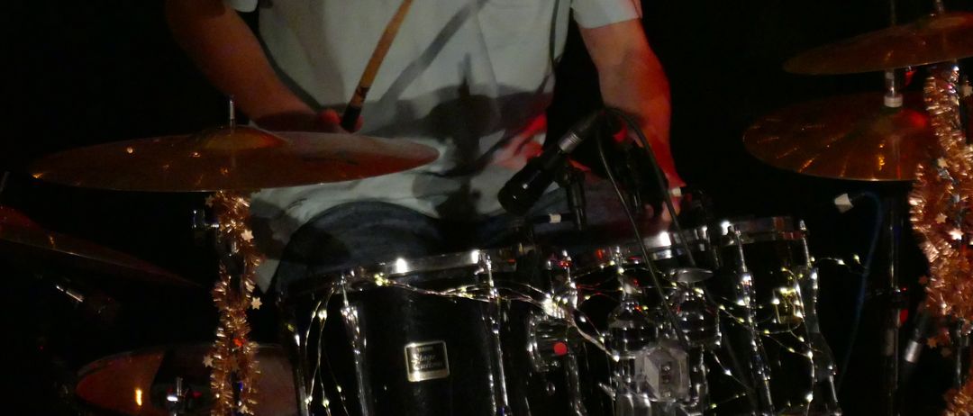 Ein Mann spielt auf der Bühne Schlagzeug, beleuchtet von Bühnenlichtern, mit Mikrofonen und Becken in der Nähe.