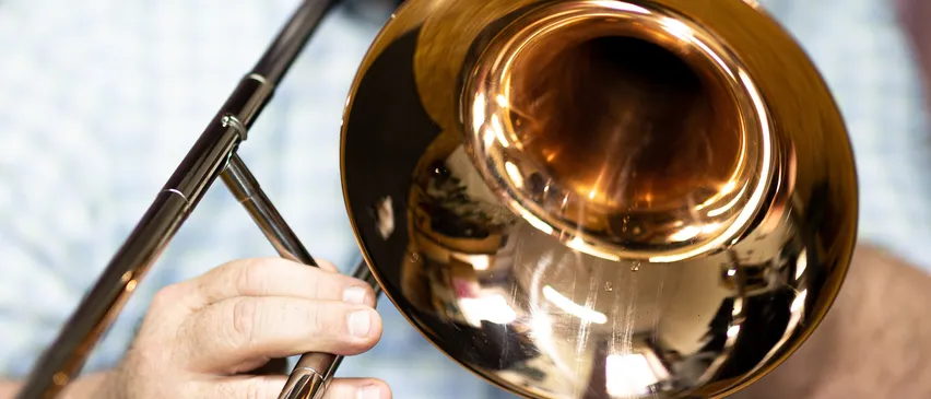 Eine Musikerhand hält ein goldglänzendes Posaune, die polierte Oberfläche reflektiert Licht. Der Posaunenmund ist leicht geöffnet, was darauf hindeutet, dass sie bereit zum Spielen ist.