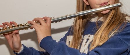 Ein junges Mädchen spielt eine silberne Flöte, konzentriert auf ihre Darbietung, mit langen Haaren und einem blauen Sweatshirt.