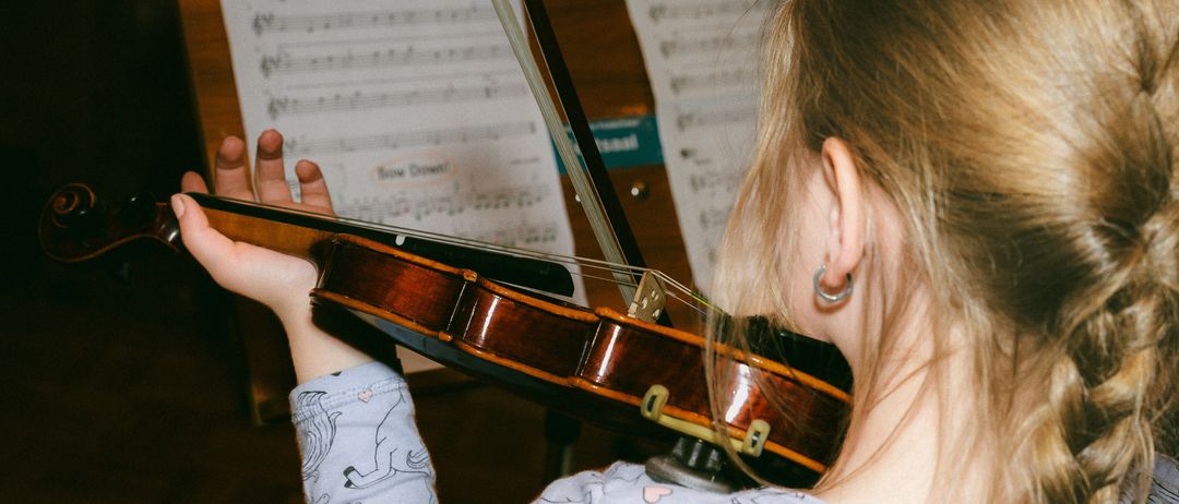 Ein Mädchen spielt Geige mit Notenblättern im Hintergrund. Die Geige ist braun mit einem schwarzen Bogen.