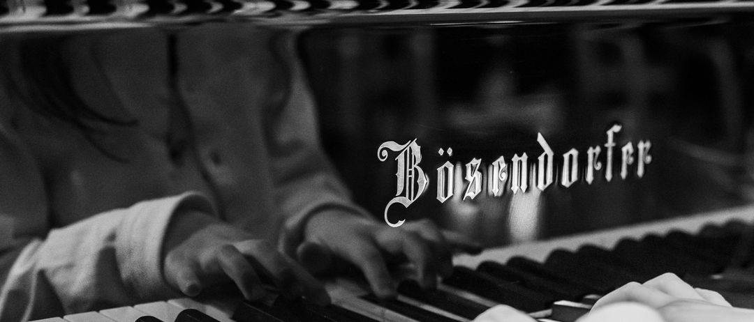 Ein Schwarz-Weiß-Bild eines Klaviers mit dem Wort 'Bosendorfer' darauf. Die Hände einer Person spielen das Klavier.