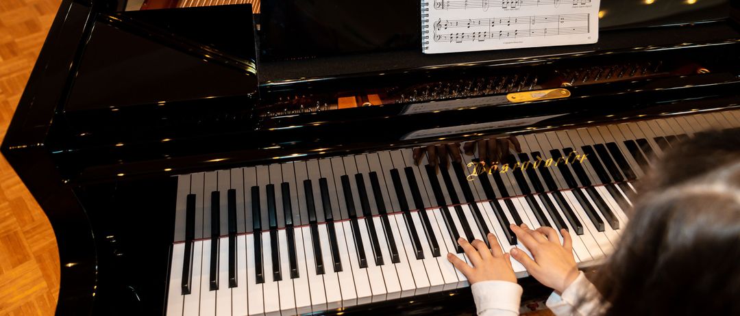 Ein junges Mädchen sitzt an einem Flügel mit einem offenen Notenheft, das eine Lokomotive zeigt. Die Klaviertasten sind sichtbar, und die Hände des Mädchens liegen auf den Tasten.