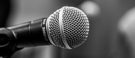 Ein Nahaufnahme eines Shure SM58 Mikrofons, schwarz und silber, mit einem Gitter. Der Hintergrund ist unscharf, wahrscheinlich in einer Aufführungsumgebung.